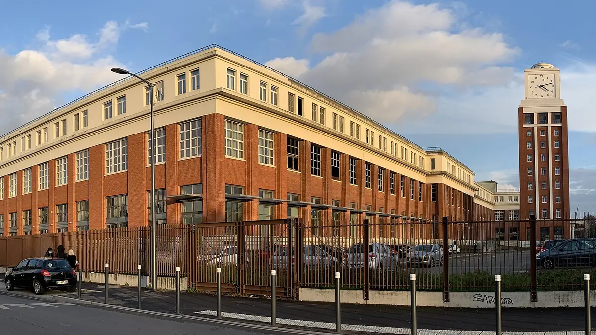 Le long bâtiment de brique et de pierre claire de l'ancienne imprimerie de L'Illustration à Bobigny, avec sa tour à horloge, derrière une grille, sous un ciel bleu.