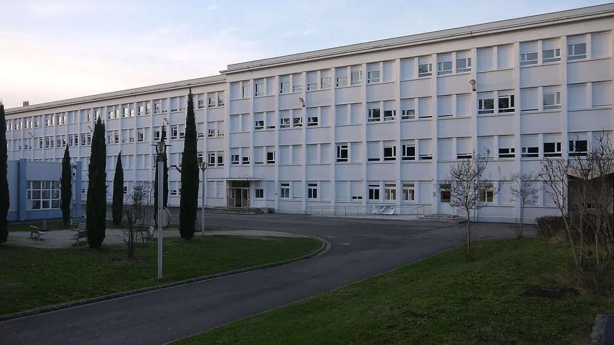Photo : la façade d'un des longs bâtiments blancs du lycée en Forêt, sur trois étages, avec une allée, une pelouse et des cyprès.