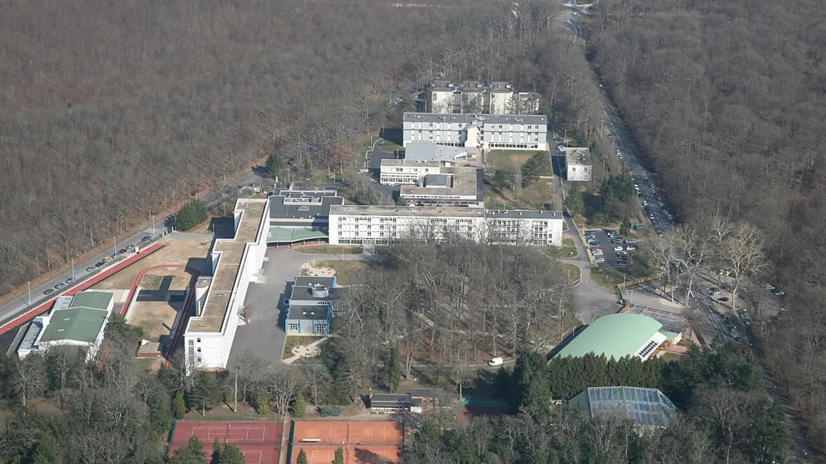 Vue aérienne du lycée en Forêt à Montargis en hiver : plusieurs bâtiments blancs, des courts de tennis, un gymnase à toit vert, entourés de tous côtés par la forêt.