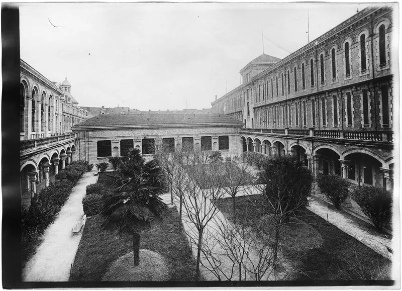 Photographie ancienne en noir et blanc : la cour d'honneur du lycée de Nîmes en 1918, un jardin planté de palmiers entouré de galeries à arcades et de bâtiments en pierre et brique.