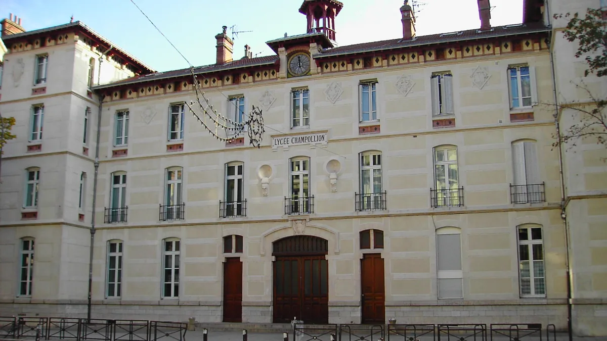 La façade du lycée Champollion à Grenoble : un long bâtiment de pierre claire à trois étages, avec une horloge sous un petit campanile, une grande porte cochère en bois et l'inscription du nom du lycée au-dessus.