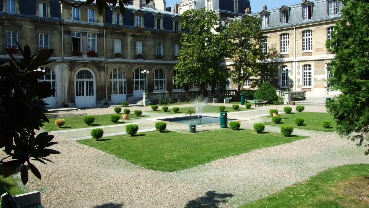 Photo : la cour d'honneur du lycée Janson-de-Sailly, pelouses et allées de gravier bordées de buissons taillés, entourées de bâtiments de pierre de la fin du XIXe siècle aux toits d'ardoise.