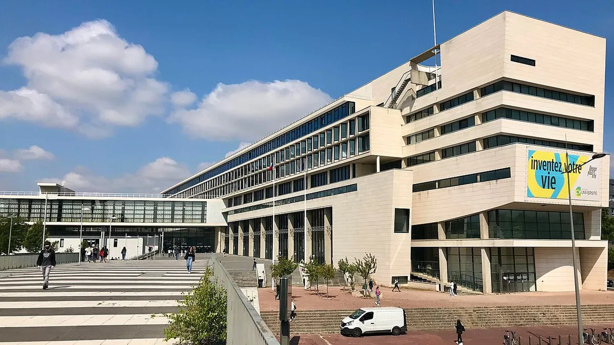 Le site des Chênes de CY Cergy Paris Université : un grand bâtiment clair aux étages en gradins et de longues façades vitrées, au bout d'un parvis où marchent des étudiants.
