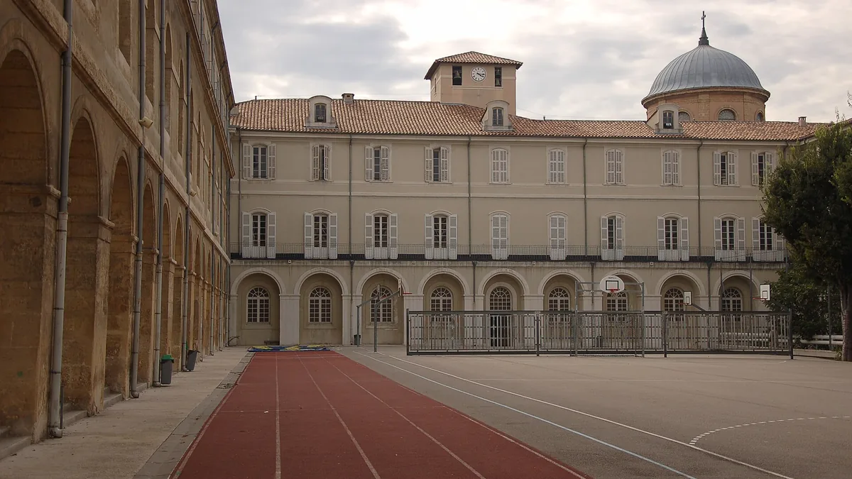 La cour du lycée Thiers : une piste d'athlétisme rouge, des terrains de basket, la façade ocre à arcades, le clocheton à horloge et, à droite, le dôme de la chapelle des Bernardines sous un ciel voilé.