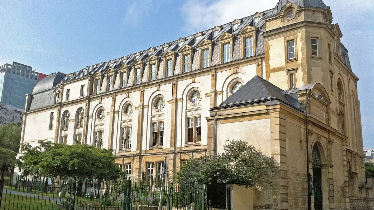 La façade du centre Vaugirard de l'université Panthéon-Assas : un bâtiment de pierre blonde du XIXe siècle avec des fenêtres rondes, un toit d'ardoise et un dôme d'angle, derrière une grille et des arbres.