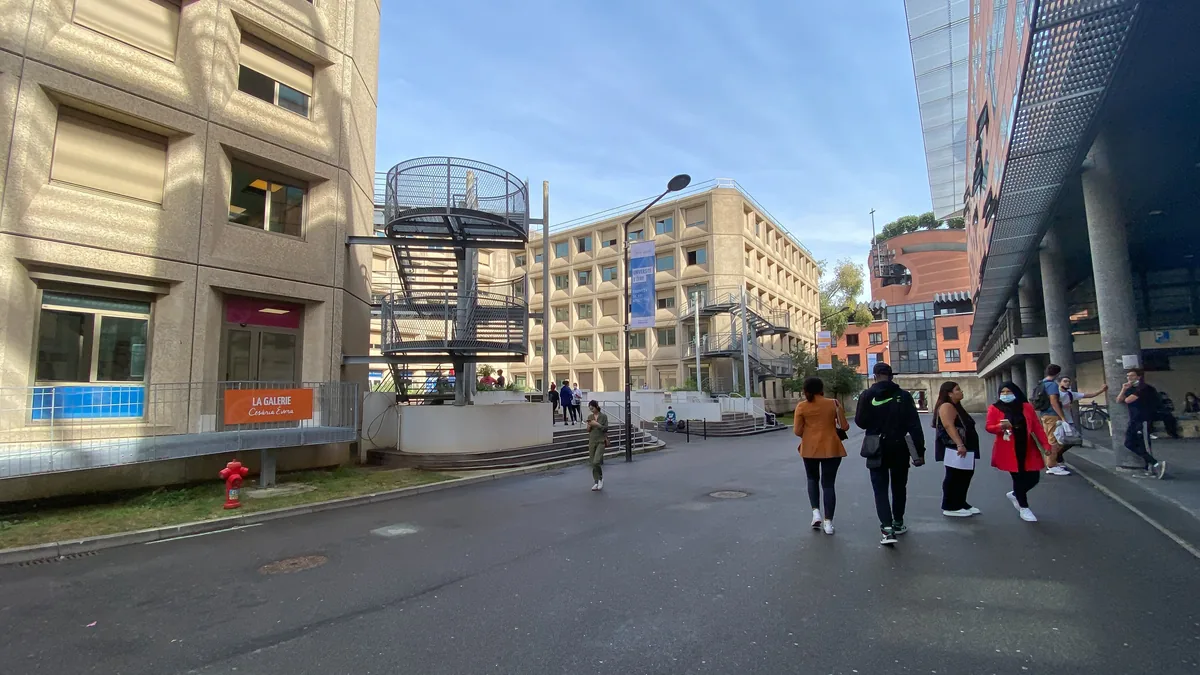 Le campus central de l'université d'Évry : des étudiants marchent entre le bâtiment Île-de-France en béton et la façade vitrée du bâtiment Maupertuis, par une matinée ensoleillée.