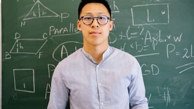 Éducateur se tenant devant un tableau noir rempli d'équations de géométrie et d'algèbre, portant des lunettes et une chemise à manches longues.