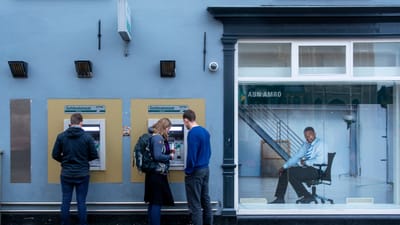 Deux DAB sur une rue de la ville avec des personnes les utilisant et une affiche de banque à proximité.