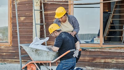 Un ouvrier du bâtiment recevant de l'aide tout en étant à genoux avec une brouette sur un chantier.