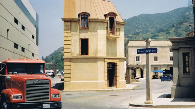 Un bâtiment rustique sur un plateau de studio de cinéma avec un camion garé près d'un fond de montagne.