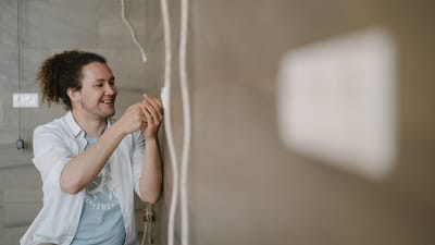 Un électricien avec des cheveux bouclés ajuste une prise à l'intérieur, souriant pendant qu'il travaille.