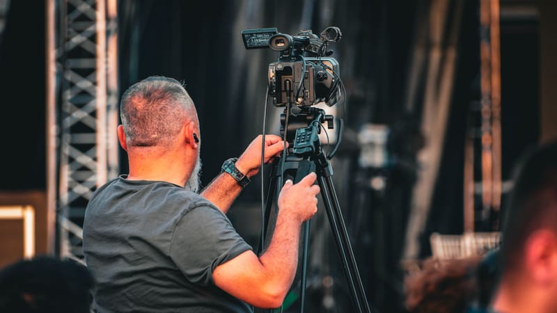 Un caméraman professionnel utilisant une caméra vidéo sur trépied lors d'un événement en plein air, capturé de derrière.