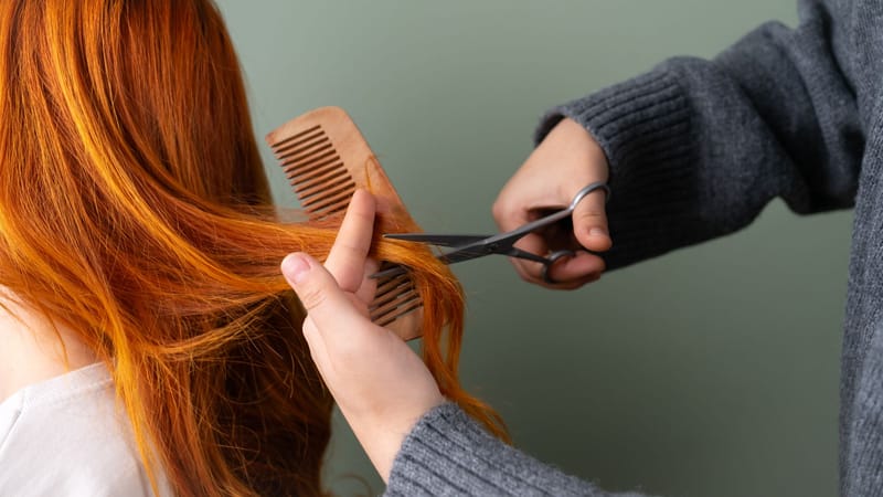 Gros plan sur des cheveux roux en cours de coupe avec des ciseaux et un peigne sur fond vert.