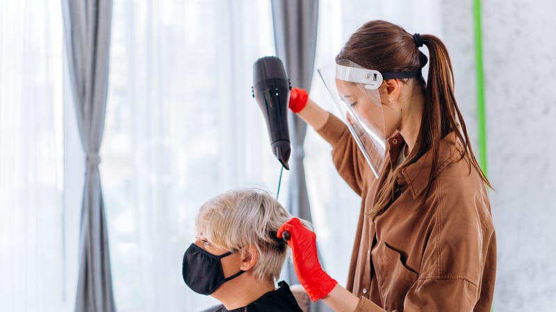 Une coiffeuse portant une visière et des gants sèche les cheveux d'un client dans un salon.