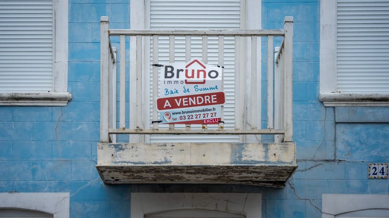 Une façade bleue usée avec un panneau 'À Vendre' sur un balcon rustique.