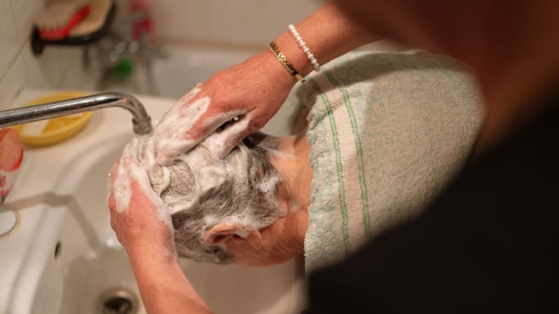 Une personne lave les cheveux d'une femme âgée dans un évier domestique, en mettant l'accent sur les soins et la toilette.