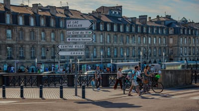 Des personnes marchant près de panneaux directionnels à Bordeaux, avec une architecture historique et des rues animées.