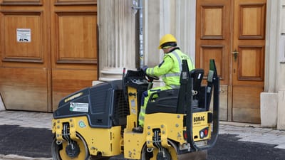Un ouvrier du bâtiment, vêtu d'un équipement de sécurité, manœuvre un rouleau compresseur sur une rue pavée près d'un bâtiment historique.