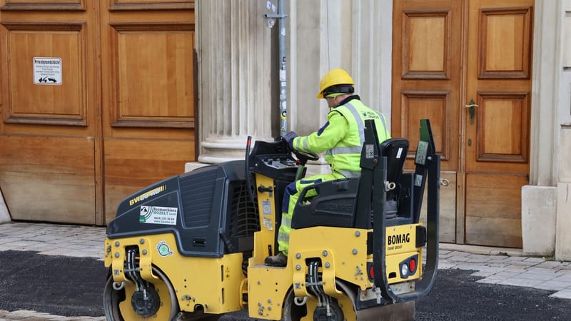 Un ouvrier du bâtiment, vêtu d'un équipement de sécurité, manœuvre un rouleau compresseur sur une rue pavée près d'un bâtiment historique.