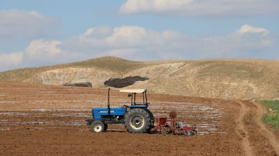 Un tracteur bleu laboure une vaste étendue de terres agricoles sur fond de collines ondulantes et de nuages.