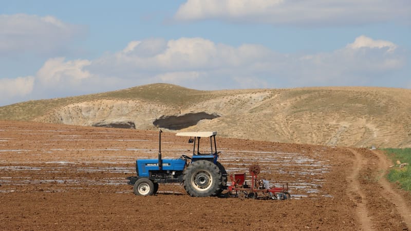 Un tracteur bleu laboure une vaste étendue de terres agricoles sur fond de collines ondulantes et de nuages.