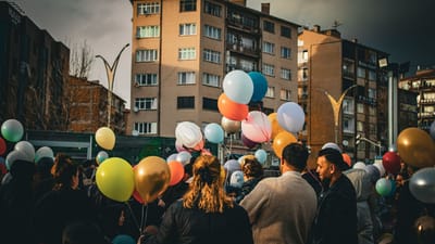 Un rassemblement vivant en plein air présente des adultes divers tenant des ballons colorés, à l'arrière-plan des immeubles sous un ciel nuageux.