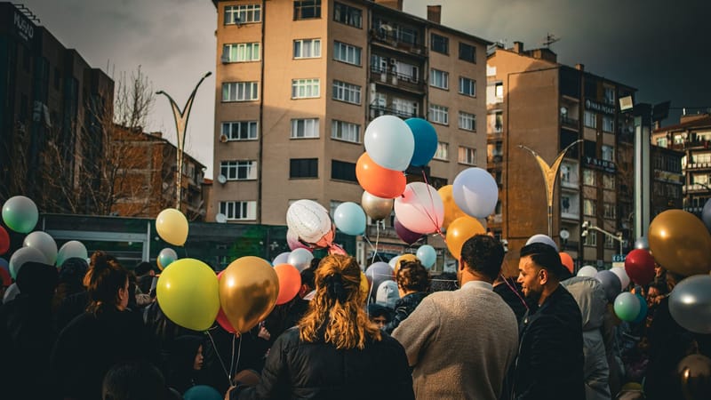 Un rassemblement vivant en plein air présente des adultes divers tenant des ballons colorés, à l'arrière-plan des immeubles sous un ciel nuageux.