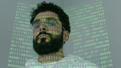 Un homme avec une barbe et des lunettes regardant la caméra, illuminé par une projection de code binaire.