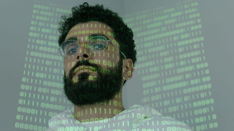 Un homme avec une barbe et des lunettes regardant la caméra, illuminé par une projection de code binaire.
