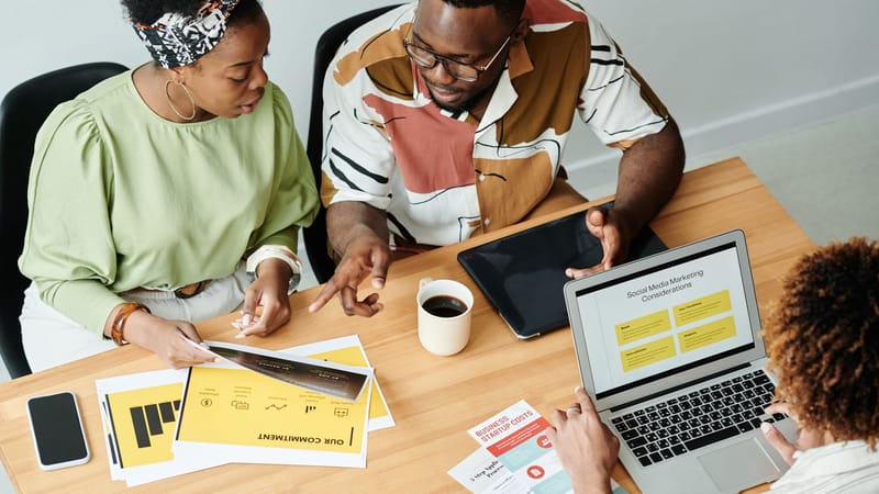 Trois collègues collaborent sur des documents marketing et un ordinateur portable dans un bureau moderne.