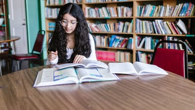 Une jeune femme étudie à une table de bibliothèque entourée de rayonnages, concentrée sur son travail.