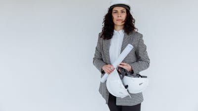 Ingénieure en tenue formelle tenant des plans et des casques de sécurité, se tenant devant un mur blanc.