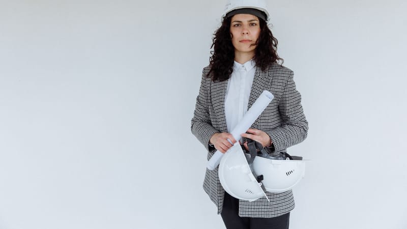 Ingénieure en tenue formelle tenant des plans et des casques de sécurité, se tenant devant un mur blanc.