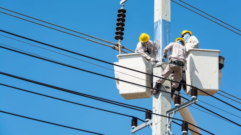 Des électriciens portant des équipements de sécurité travaillent sur des lignes électriques à l'aide d'élévateurs sous un ciel bleu dégagé.