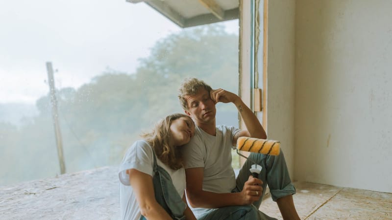 Un couple prend une pause pendant la rénovation de leur maison, assis à l'intérieur avec un rouleau à peinture.