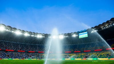 Vue nocturne d'un stade de football avec des arroseurs arrosant le terrain, renforçant l'atmosphère dynamique.