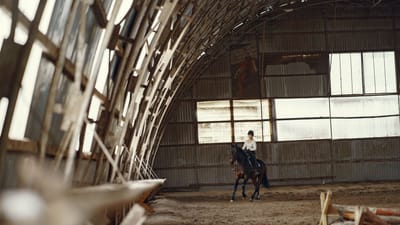 Une femme monte un cheval dans une salle de manège rustique entourée de structures en bois.