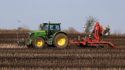 Un tracteur vert avec une herse rouge laboure une terre brune sous un ciel bleu clair.