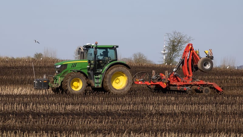 Un tracteur vert avec une herse rouge laboure une terre brune sous un ciel bleu clair.