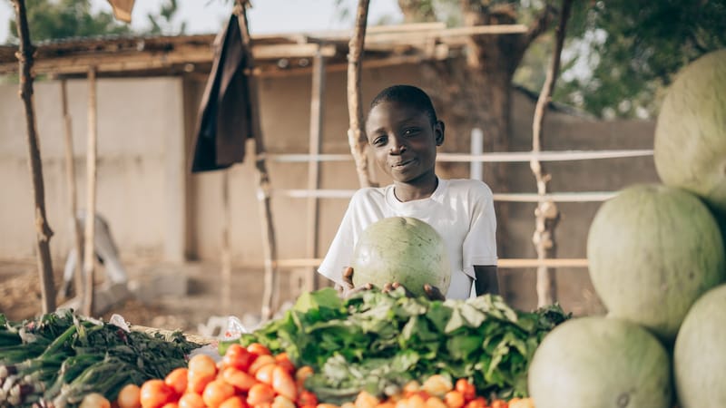 Un jeune garçon avec des légumes frais et des pastèques sur un stand de marché en plein air.