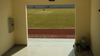 Vue ensoleillée depuis un couloir intérieur, donnant sur un terrain de football en herbe avec une personne au travail.