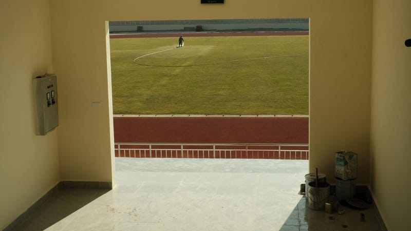 Vue ensoleillée depuis un couloir intérieur, donnant sur un terrain de football en herbe avec une personne au travail.