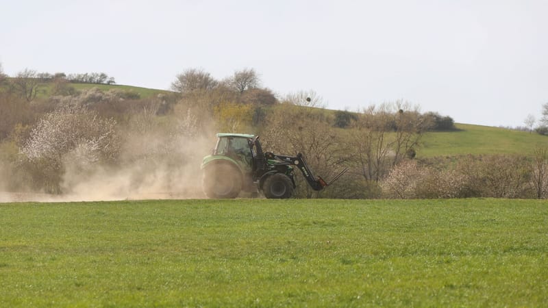 Un tracteur laboure un champ ensoleillé au printemps, créant un nuage de poussière sur un paysage rural.