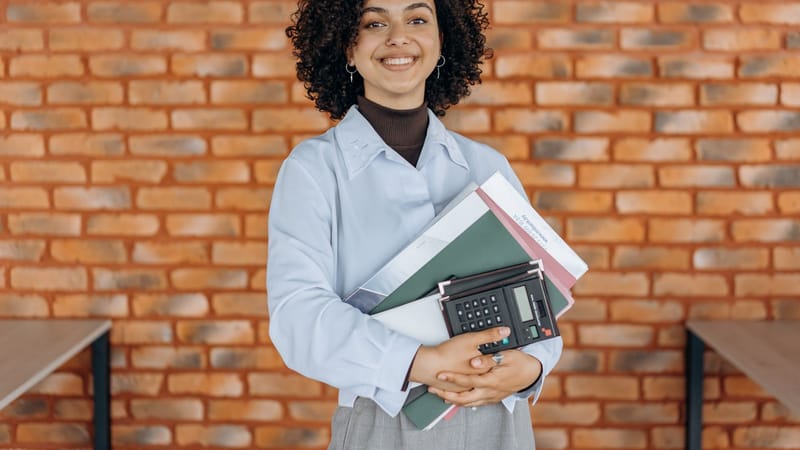 Femme joyeuse aux cheveux bouclés tenant des dossiers et une calculatrice devant un mur en briques.