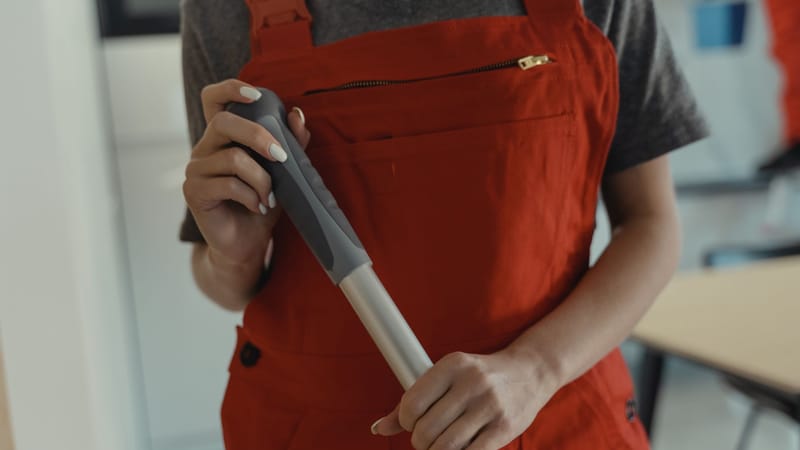 Une femme en tablier rouge tenant un manche à balai, en train de nettoyer à l'intérieur.