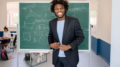 Un enseignant souriant se tient à côté du tableau noir dans une salle de classe, illustrant des concepts mathématiques.