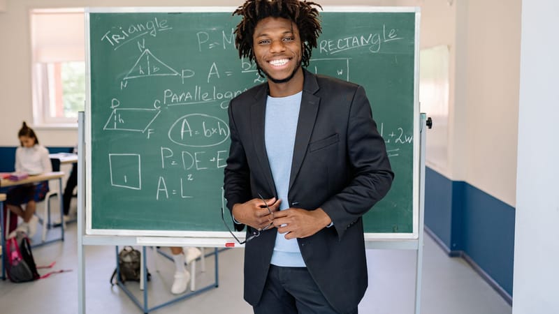 Un enseignant souriant se tient à côté du tableau noir dans une salle de classe, illustrant des concepts mathématiques.