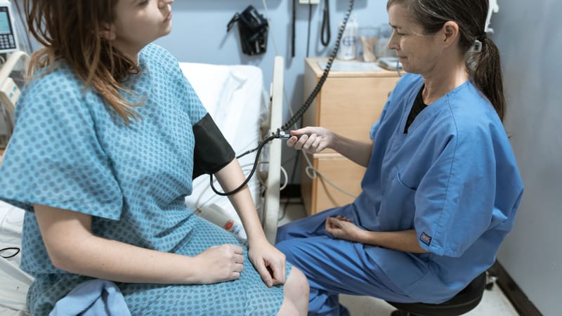 Une infirmière en tenue médicale bleue vérifie la tension artérielle d'une patiente dans une chambre d'hôpital.