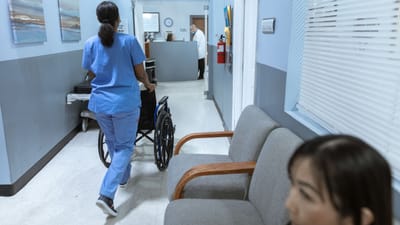 Une infirmière en tenue médicale pousse un fauteuil roulant à travers un couloir d'hôpital bien éclairé, s'occupant des soins aux patients.