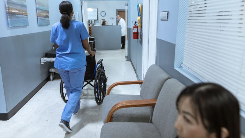 Une infirmière en tenue médicale pousse un fauteuil roulant à travers un couloir d'hôpital bien éclairé, s'occupant des soins aux patients.
